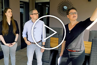 Drei Personen in einer Terrassensituation, eine Person zeigt auf etwas, im Hintergrund eine runde Wanddekoration.