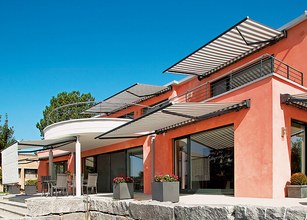 Moderne Architektur mit Terrassensituation, Markisen und durchgehender Glasfront vor blauem Himmel. 