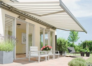 Markise in hellen Streifen über Terrasse, umgeben von Pflanzen und modernen Möbeln. 