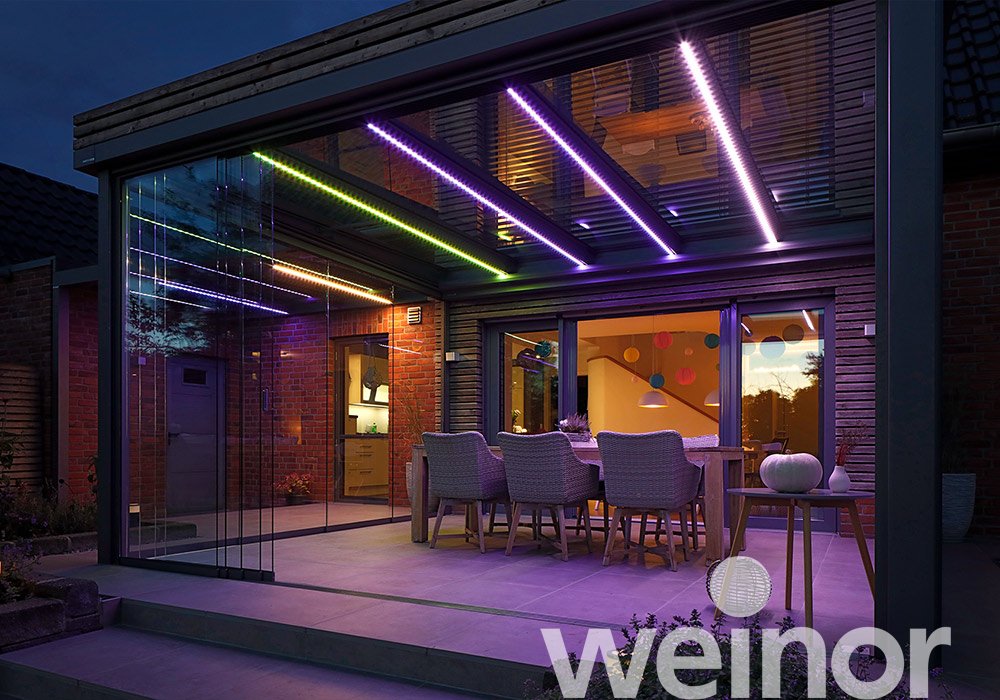 Moderne Überdachung mit Glas-Schiebewänden und bunten LED-Lichtern in einer Terrassensituation bei Nacht.