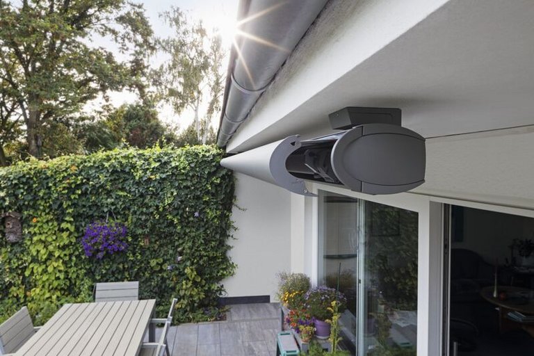 Markisenkasten und Terrassensituation mit einem grünen Sichtschutz und Pflanzen im Hintergrund.