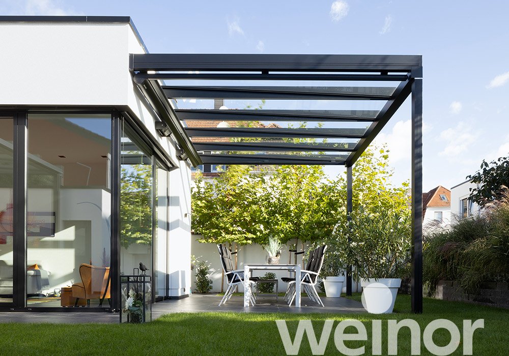 Moderne Überdachung mit Glas und einem Tisch sowie Stühlen in einer Terrassensituation. Dominante Farben: Schwarz und Grün.