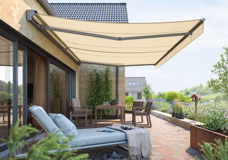 A terrace with a beige awning extending over seating and a sun lounger, surrounded by greenery in a bright setting.