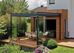 Terrassensituation mit Überdachung, Glas-Schiebewänden und Holzverkleidung, umgeben von Grünpflanzen und Rasen. 