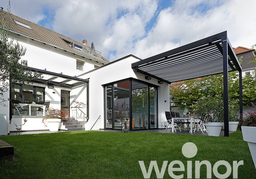 Haus mit Überdachung und Glasflächen, umgeben von Rasen und Pflanzen, unter einem bewölkten Himmel.