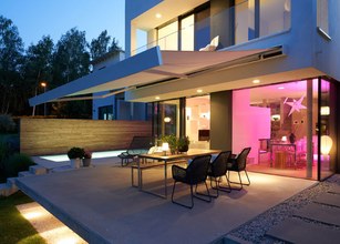 Moderne Terrasse mit Überdachung, Stühlen und Tisch, nachts beleuchtet mit warmen Farben. 