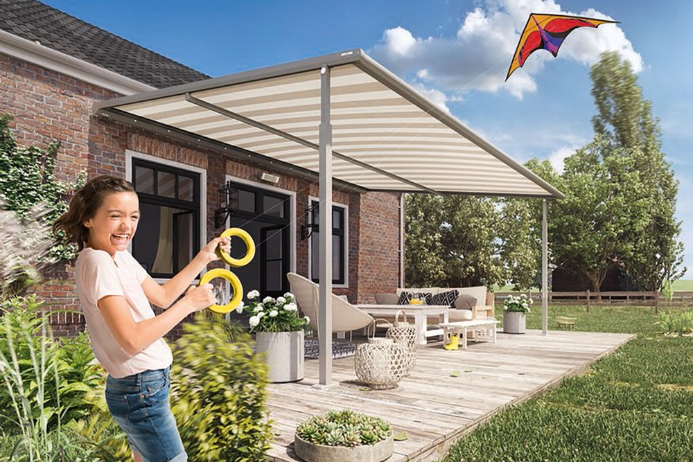 Enfant jouant avec un cerf-volant sous une pergola, sur une terrasse en bois, avec des plantes autour.