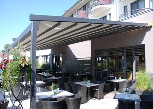 Terrassensituation unter einer Überdachung mit Markisenkasten, umgeben von Pflanzen und Tischen. 