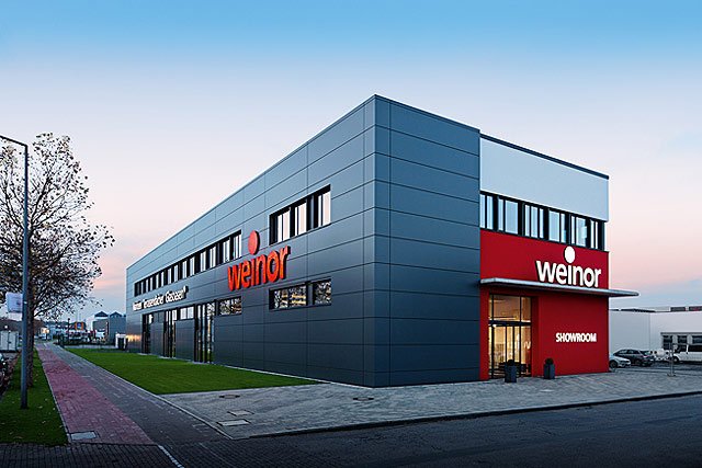 Moderner, rechteckiger Gebäudekomplex in Grau mit roten Akzenten und großen Fenstern. Moderner, rechteckiger Gebäudekomplex in Grau mit roten Akzenten und großen Fenstern.
