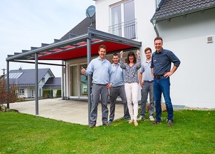 Gruppe von fünf Personen, die lächelnd vor einer Überdachung mit rotem Markisentuch steht. 