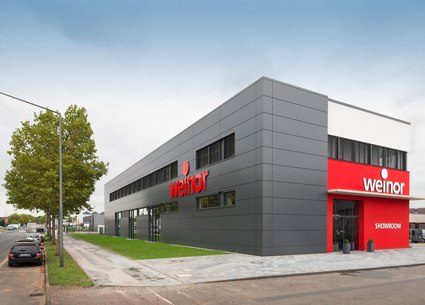 Bâtiment moderne avec des murs gris et une enseigne rouge, entouré de verdure.