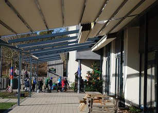 Markisen und Überdachung über einem Gehweg, Kinder in bunten Kleidern gehen vorbei, Weihnachtsbaum sichtbar. 