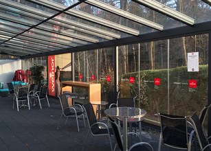 Terrassensituation mit Glas-Schiebewänden, Markisenkasten und Tischen mit Stühlen. Dunkles Holz und klare Glasflächen. 