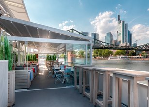 Terrassensituation mit Überdachung, Blick auf Gewässer und Stadtlandschaft, helle Farben und moderne Architektur. 