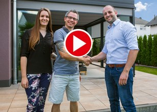 Drei Personen schütteln sich die Hände vor einem modernen Glasbau mit Terrassensituation im Hintergrund. 