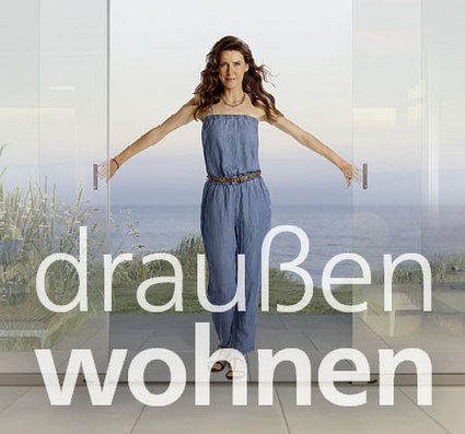 Vrouw met bruin haar in een blauwe jumpsuit, staand tussen glazen deuren met uitzicht op de zee.