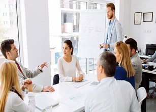 Menschen in einem modernen Büro, die an einem Tisch sitzen und einem Präsentierenden zuzuhören. 