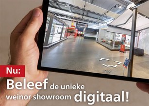 Een hand houdt een tablet met een digitale weergave van een showroom. Achtergrond is neutraal en lichtgrijs. 