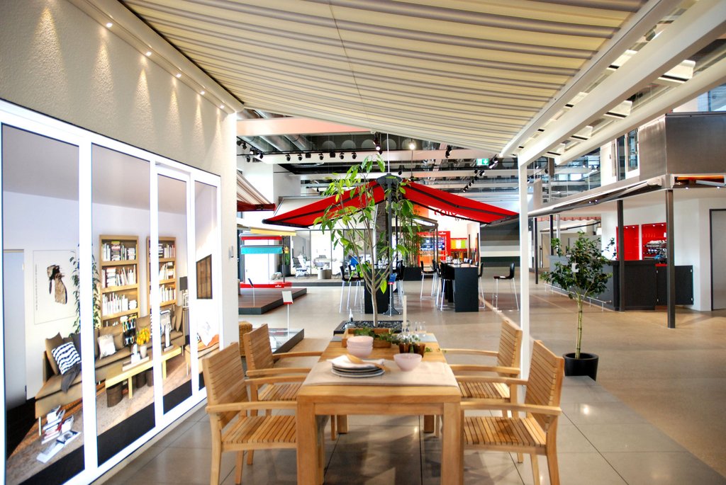 Terrassensituation mit einem Esstisch und roten Überdachungen im Innenraum einer Ausstellung. Terrassensituation mit einem Esstisch und roten Überdachungen im Innenraum einer Ausstellung.