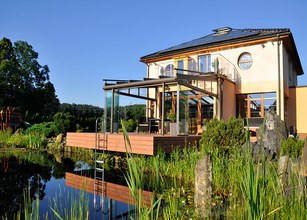 Moderne Hausfassade mit Wintergarten, Terrasse, Wasseransicht und umliegender Bepflanzung. 