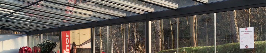 Eine moderne Überdachung mit Glas-Schiebewänden und Sonnenlicht, im Hintergrund ein bewaldeter Bereich. Eine moderne Überdachung mit Glas-Schiebewänden und Sonnenlicht, im Hintergrund ein bewaldeter Bereich.