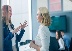 Zwei Frauen unterhalten sich in einem Büro, eine trägt einen blauen Blazer, die andere ein weißes Hemd. 