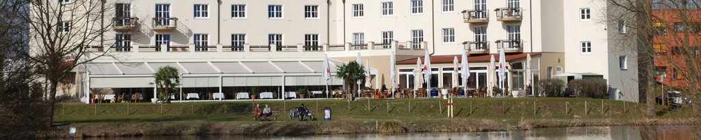 Building facade with large windows, balconies and a terrace near a water body, featuring greenery in the foreground. Building facade with large windows, balconies and a terrace near a water body, featuring greenery in the foreground.