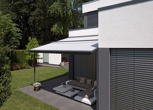 Moderne Überdachung in grau mit Markisentuch, daneben grüne Terrassensituation und helles Wohnhaus. 