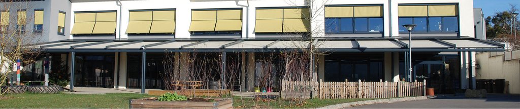 Moderne gebouw met gele luifels en overkapping. Groen gras en houten hekwerk zichtbaar.