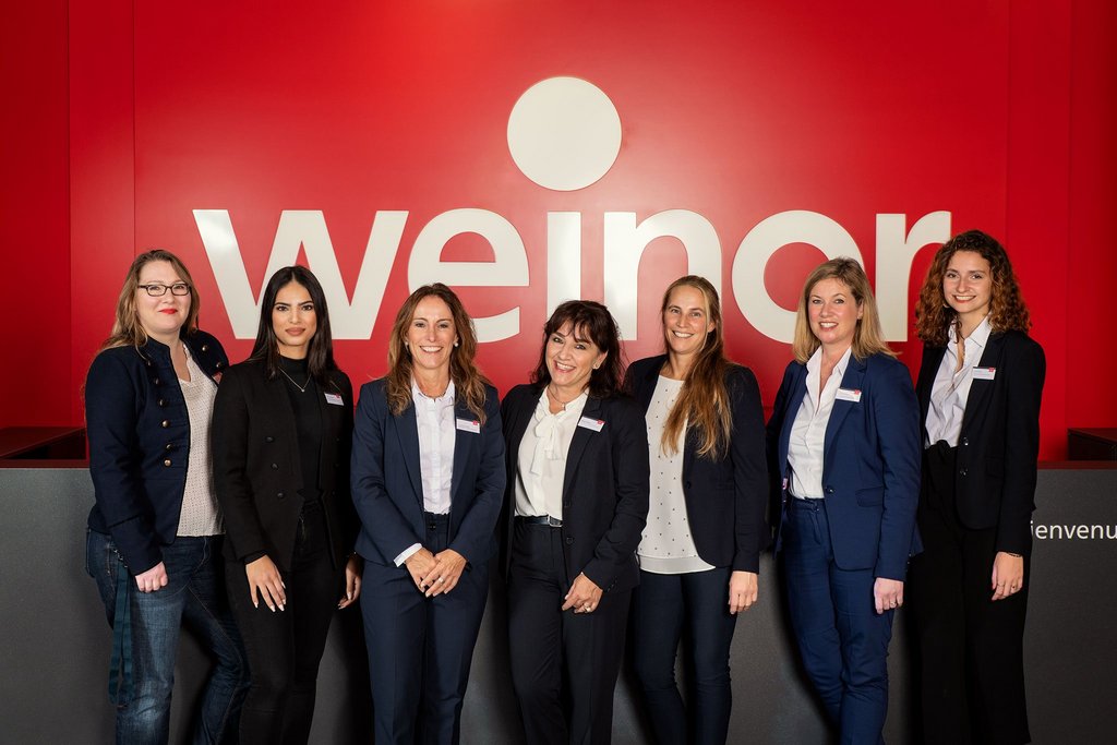 Groupe de femmes souriantes en tenue professionnelle devant un fond rouge avec le logo weinor. Groupe de femmes souriantes en tenue professionnelle devant un fond rouge avec le logo weinor.