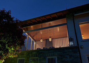 Überdachung mit Beleuchtung und Glas-Schiebewänden, im Hintergrund ein Baum, nächtliche Atmosphäre. 