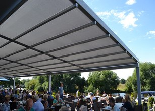 Überdachung mit Grautönen, darunter zahlreiche Personen in einer Terrassensituation am Wasser. 