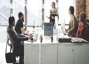 Fünf Personen im modernen Büro präsentieren und diskutieren, umgeben von großen Fenstern und hellem Licht. 